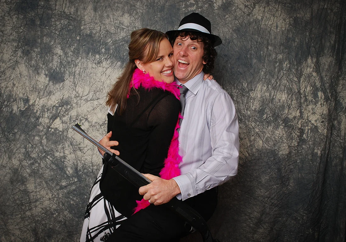 Couple hugging with a pink feather boa, black hat, and a toy gun.