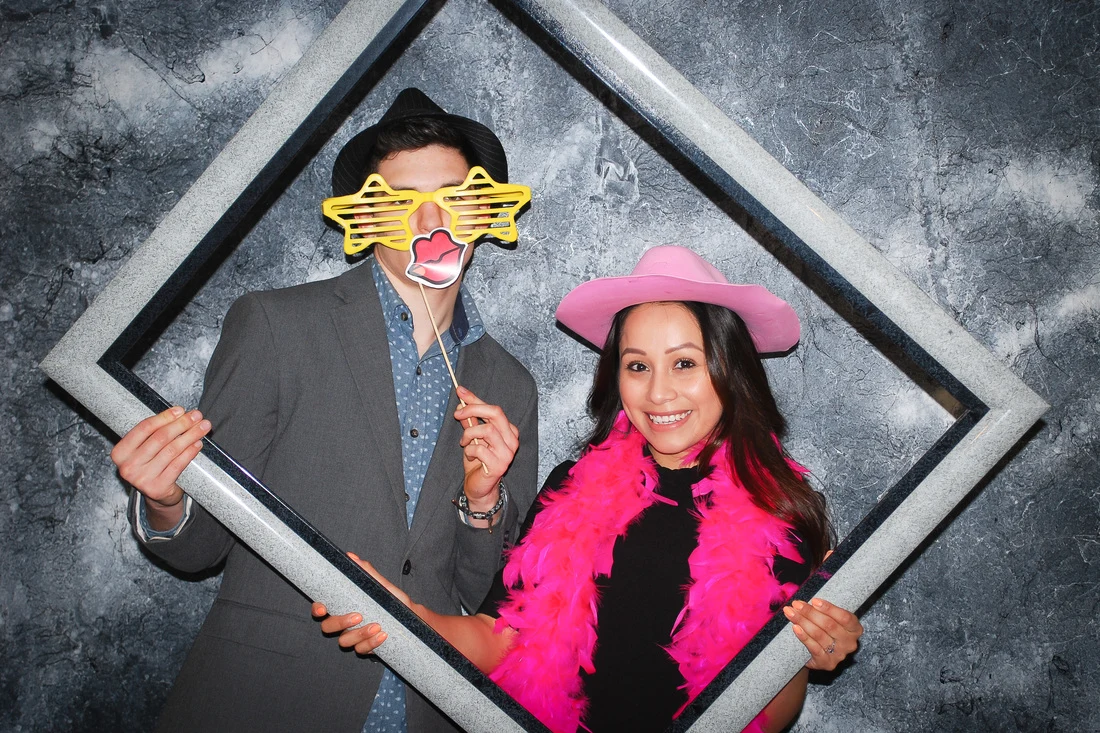 Two people holding a large picture frame, wearing star glasses, a pink cowboy hat, and a pink boa.