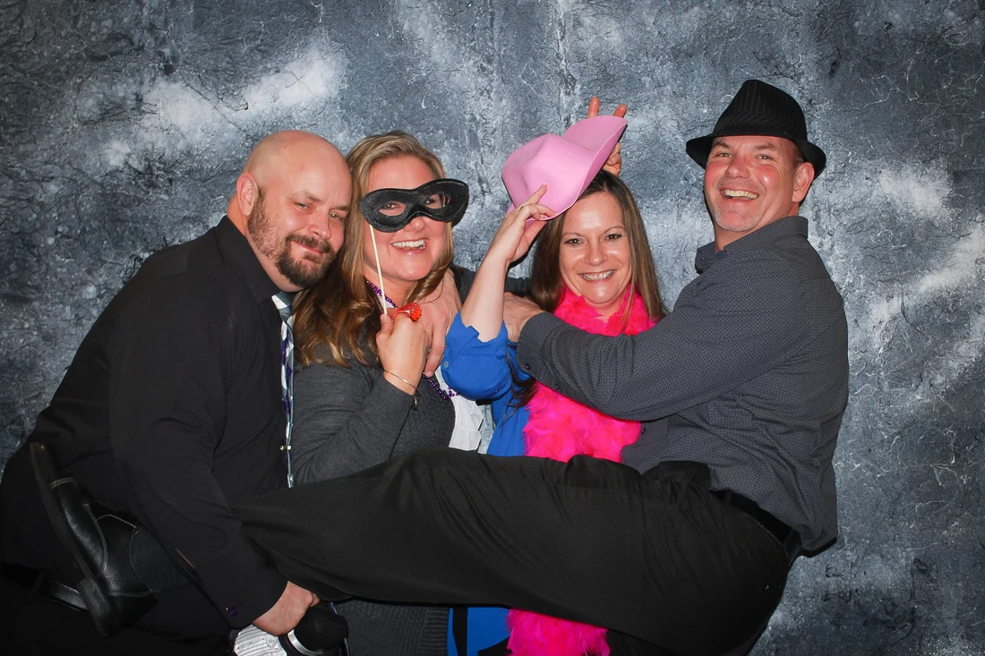 Three people holding a fourth person sideways while posing with hats, a black mask, and a pink boa.