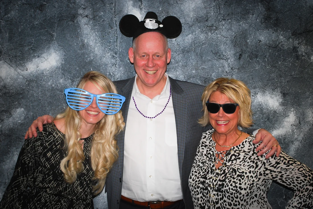 Three smiling people wearing party props, including mouse ears, oversized blue glasses, and sunglasses.