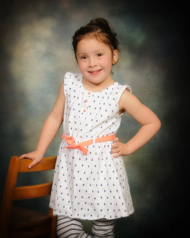 Child in a white patterned dress with a coral belt posing beside a wooden chair.