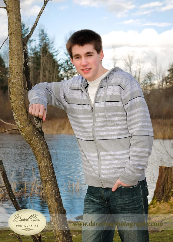 Person in a gray striped sweater standing beside a tree at the water's edge.