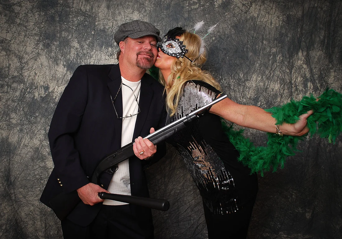 Woman in a decorative mask kissing a man in a gray cap, with a green feather boa and a toy gun.