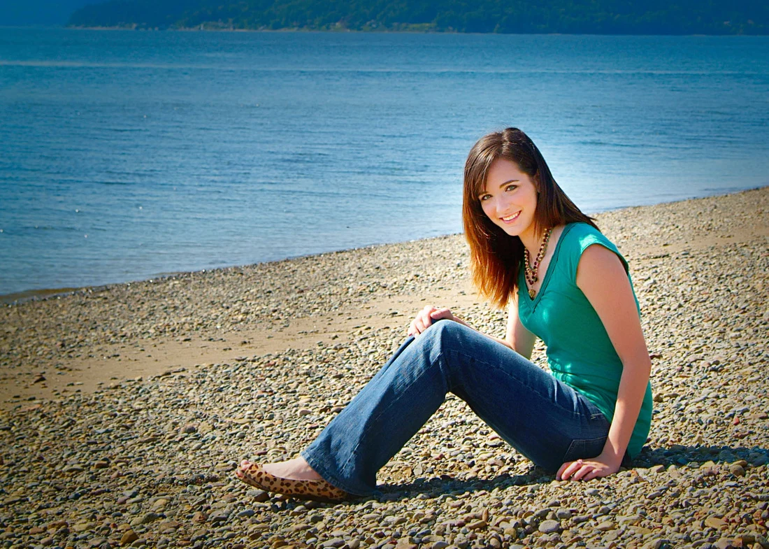 Person in a turquoise top and jeans sitting on a pebbled shore beside blue water.