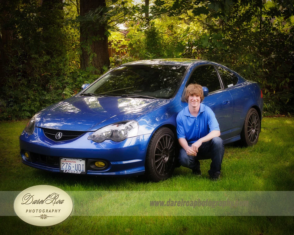 Person in a blue polo shirt crouching beside a blue car in a wooded yard.