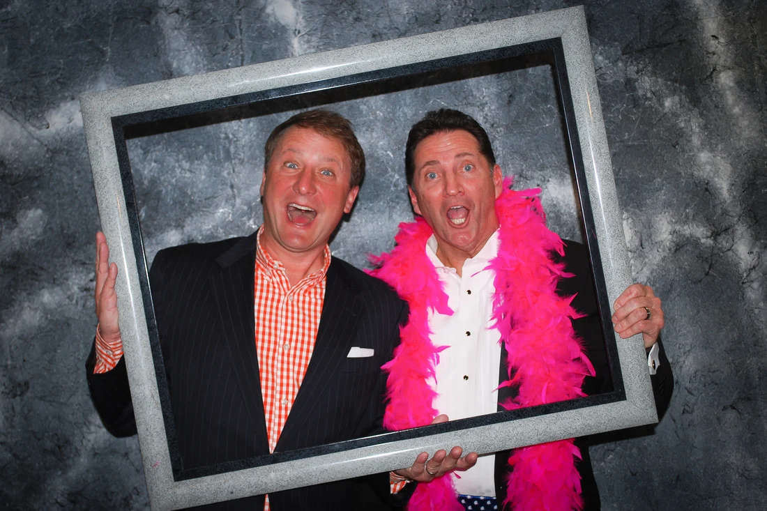 Two men making surprised expressions inside a large picture frame, one wearing a bright-pink feather boa.