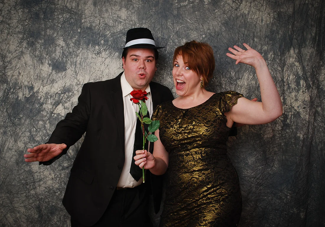 Man in a black hat and woman in a gold-and-black dress making playful faces while holding a red rose.