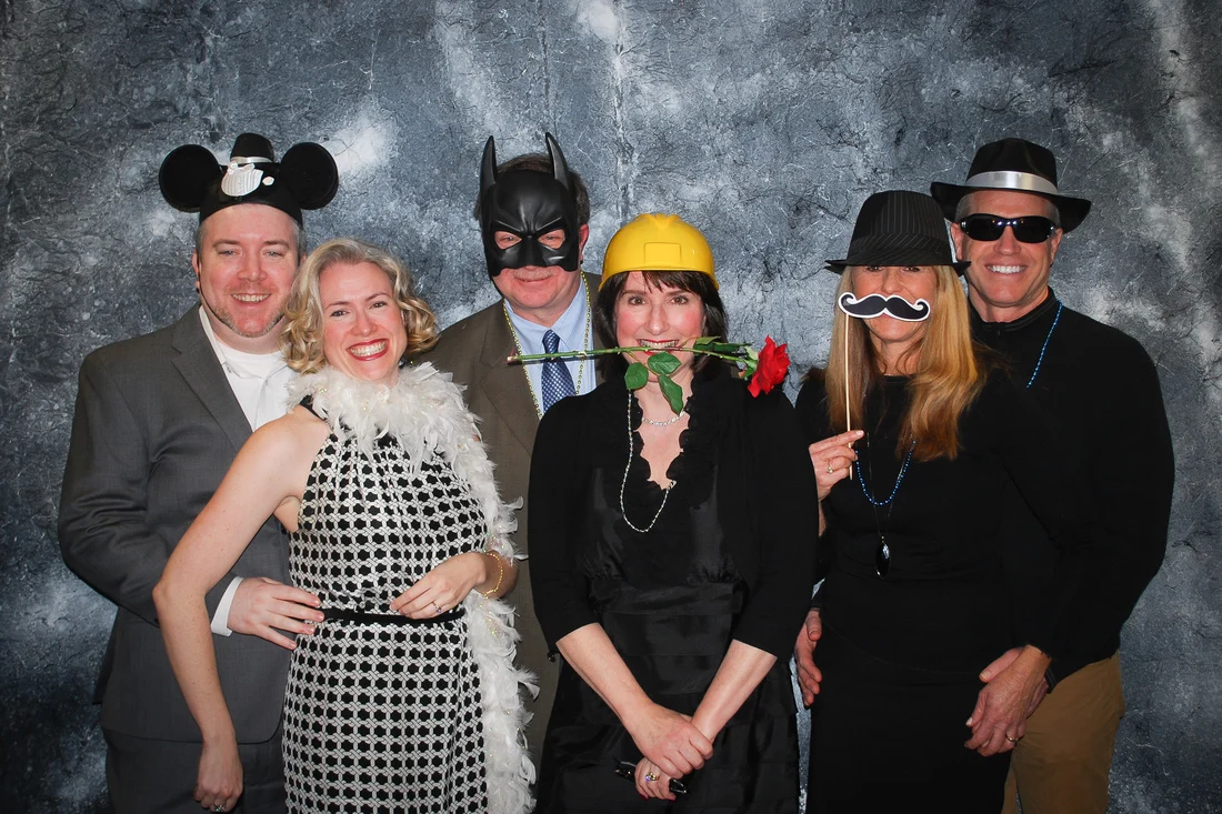 Six people posing with mouse ears, a black mask, hats, a toy mustache, a white boa, and a rose.