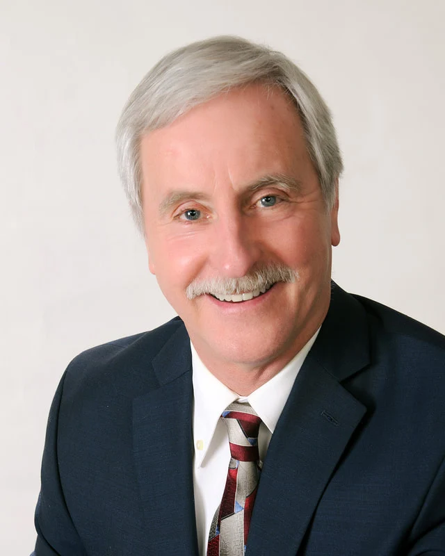 Smiling man with gray hair and a mustache wearing a navy suit and striped tie against a pale backdrop.