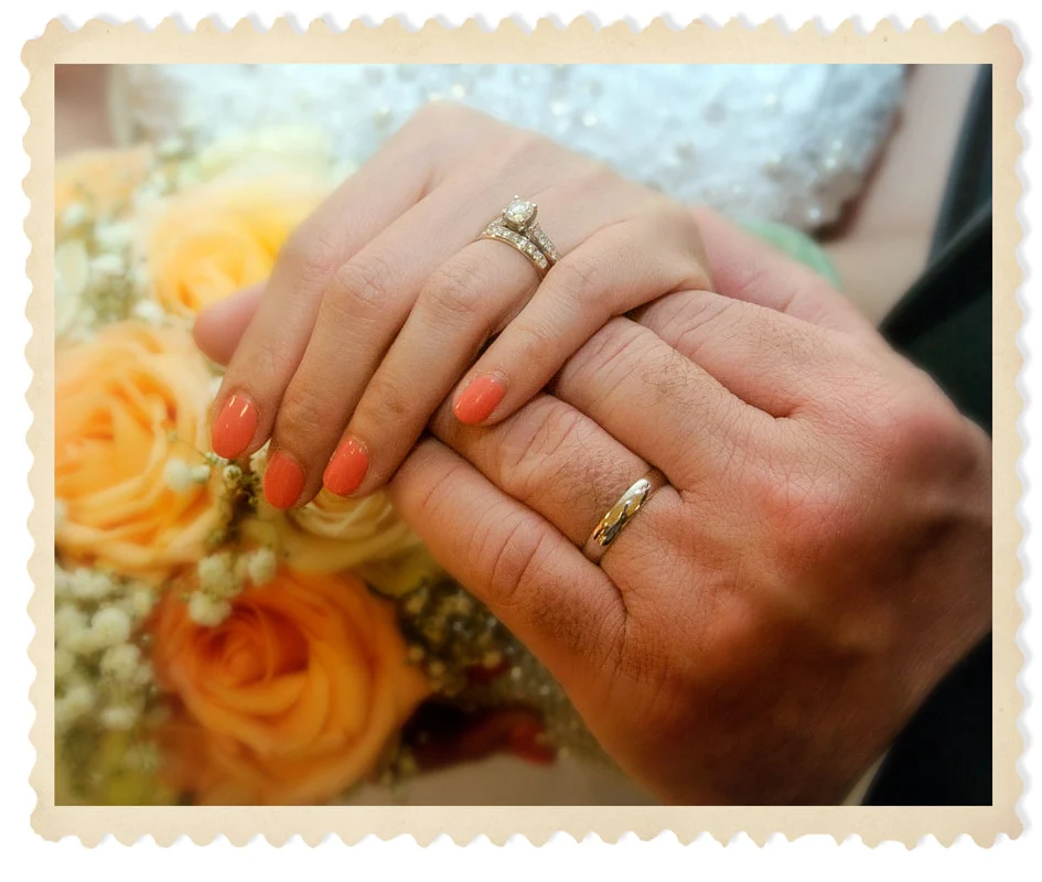 Close-up of two hands wearing wedding rings beside a bouquet of peach roses.