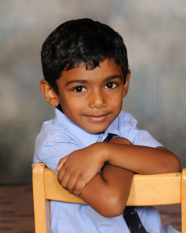 Child in a light-blue shirt and tie resting folded arms on the back of a wooden chair.