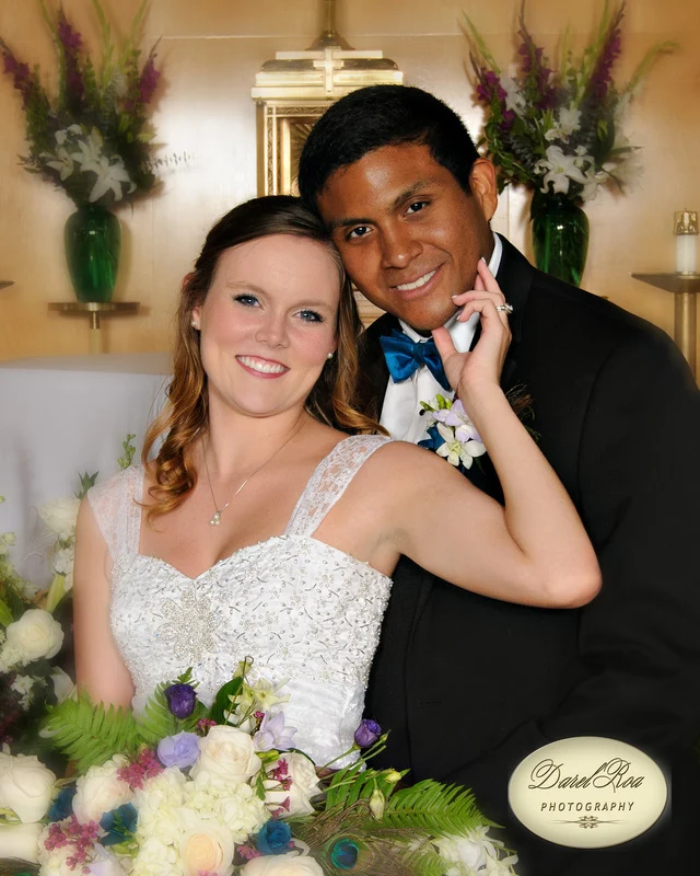 Smiling bride holding a bouquet and touching the groom's cheek in front of floral arrangements.