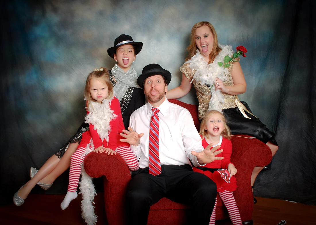 Two adults and three children making playful faces around a red sofa, with hats, feather boas, and red outfits.