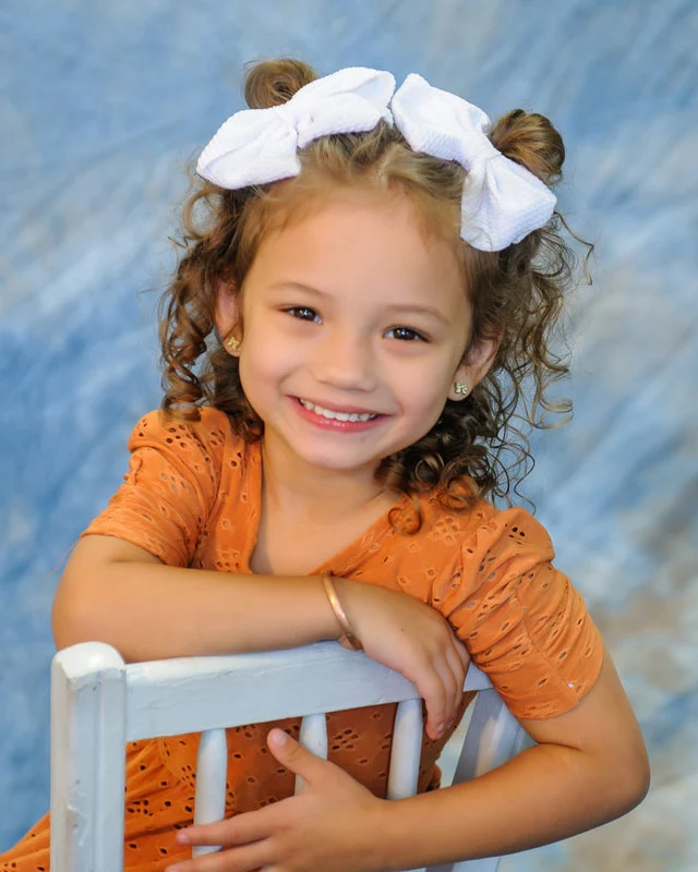 Smiling child with white hair bows and an orange top leaning on a white chair.
