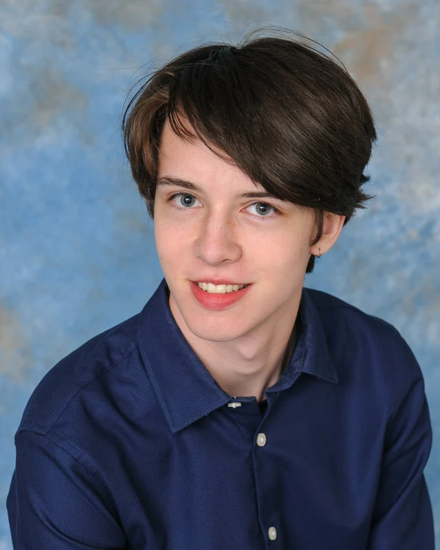 Smiling teenager in a dark-blue button-up shirt against a blue studio backdrop.