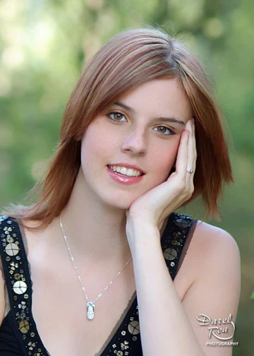 Person with shoulder-length hair resting their cheek on one hand against a leafy green background.