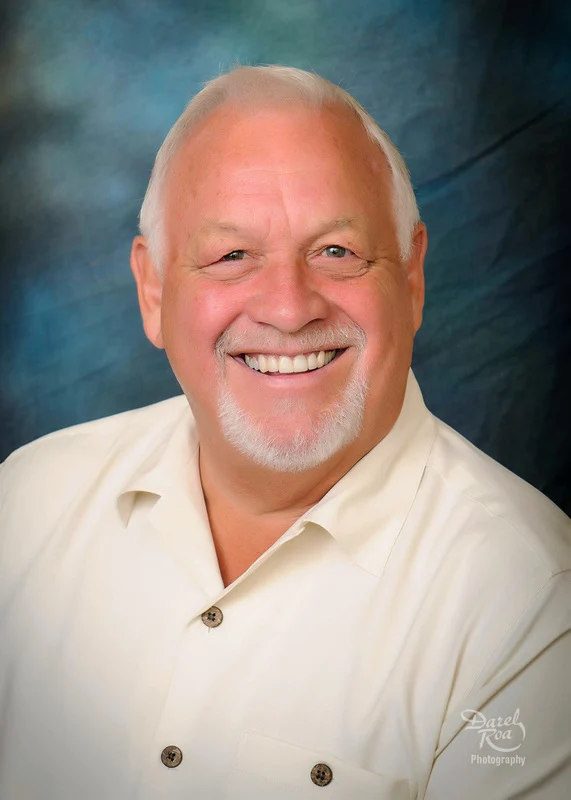 Smiling man with white hair and a white goatee wearing a cream shirt against a blue backdrop.