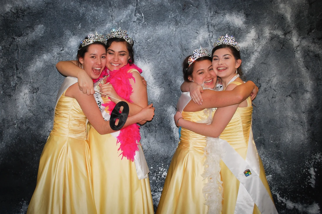 Four people in yellow gowns and tiaras hugging in pairs, with a bright-pink feather boa.
