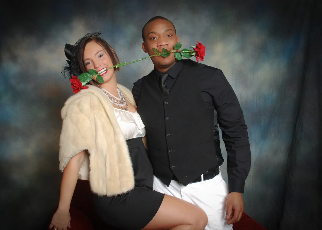 Two people posing in party clothes with red roses held in their mouths.