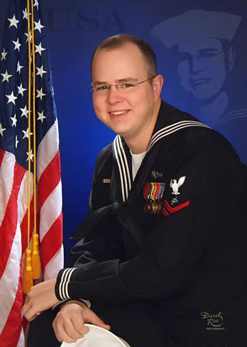 Man in a sailor uniform sitting beside an American flag and holding a white cap.