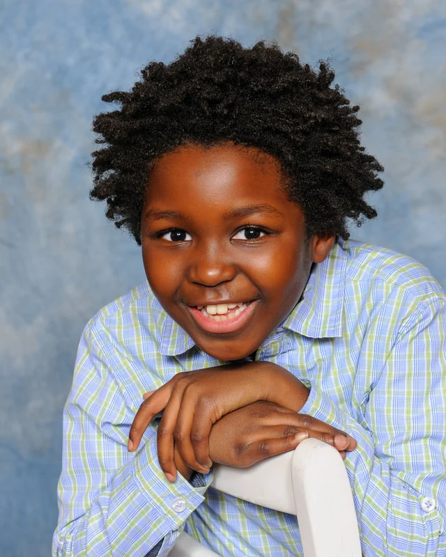 Smiling child in a blue-and-green checked shirt leaning on a white chair against a blue backdrop.