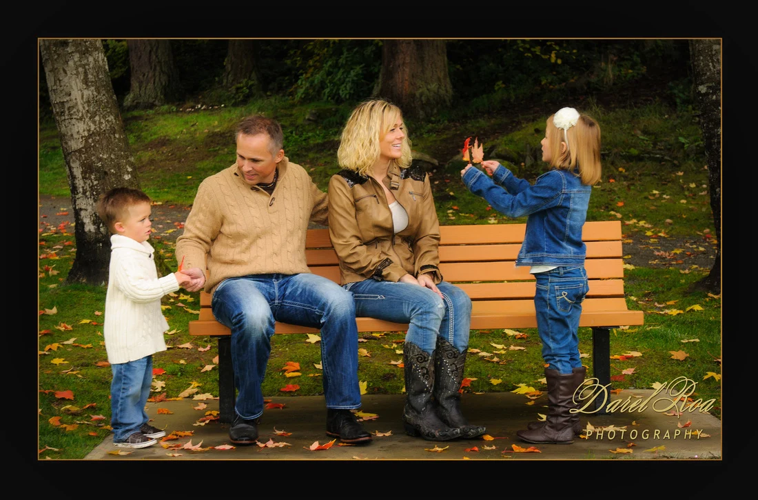 Two adults sitting on a park bench while two children stand beside them among autumn leaves.