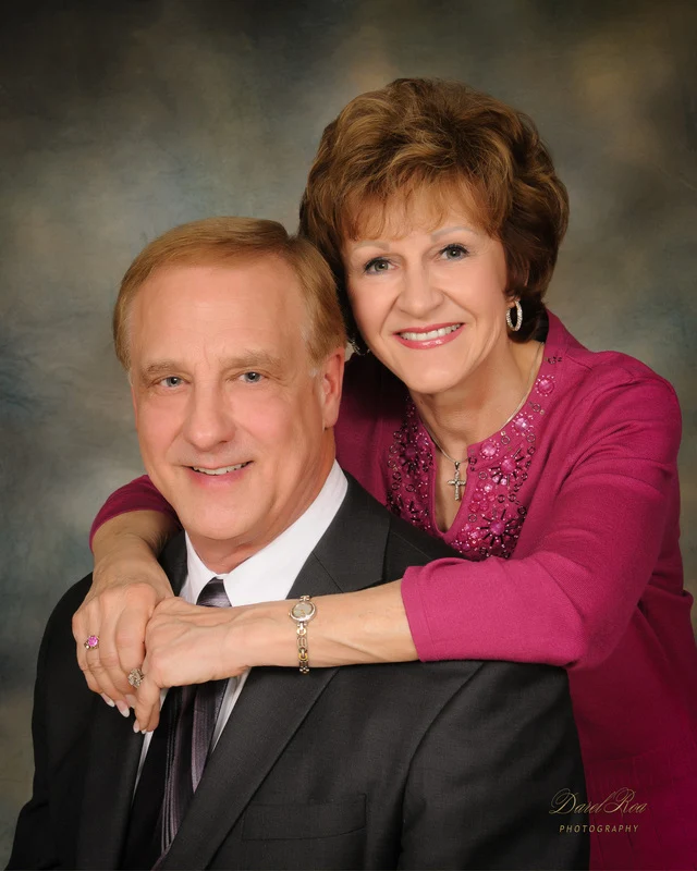 Smiling woman in a magenta top resting her arms around a seated man in a dark suit.