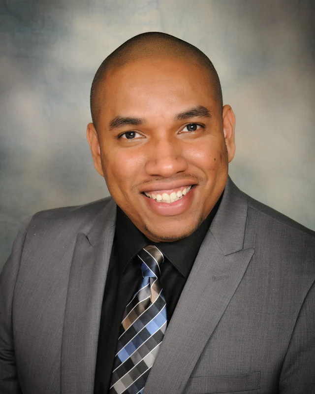 Smiling man in a gray suit, black shirt, and patterned tie against a gray studio backdrop.