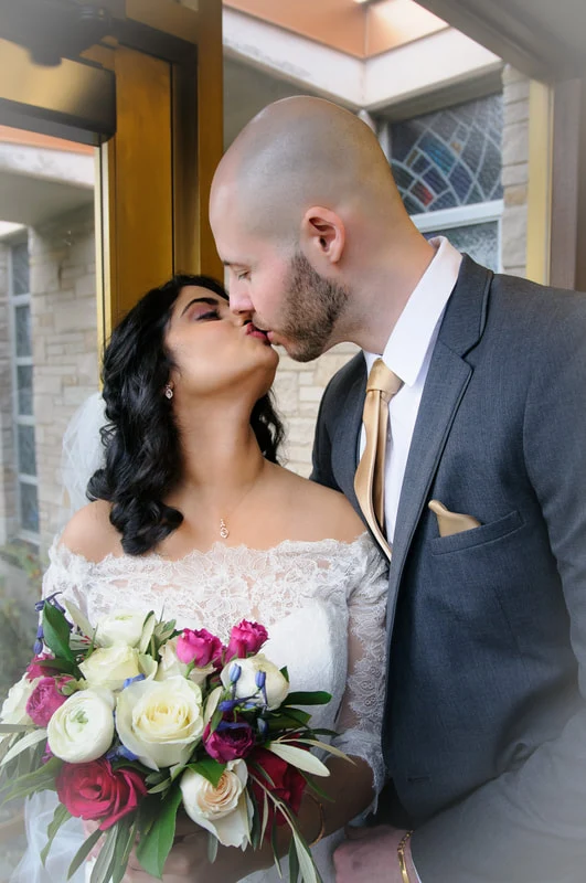 Bride holding a colorful bouquet kisses a groom in a gray suit beside a doorway.