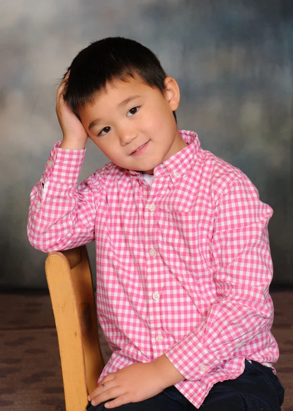 Child in a pink checked shirt sitting on a wooden chair with one hand against their head.