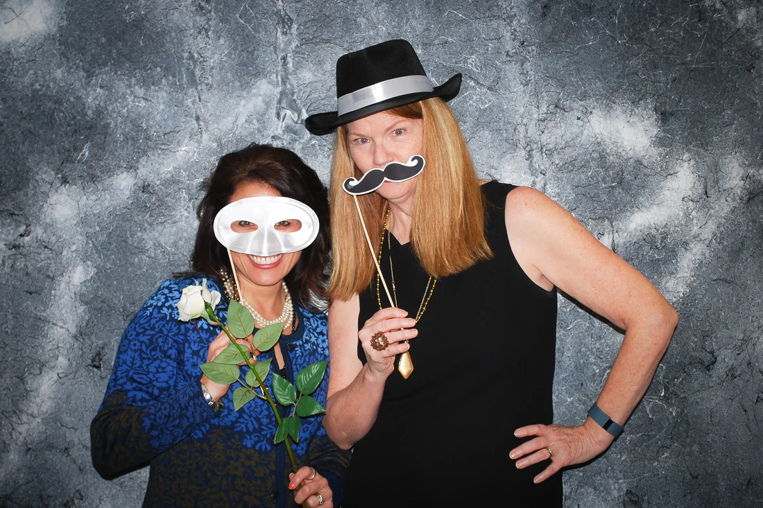 Two women posing with a white mask, a toy mustache, a black hat, and a white rose.