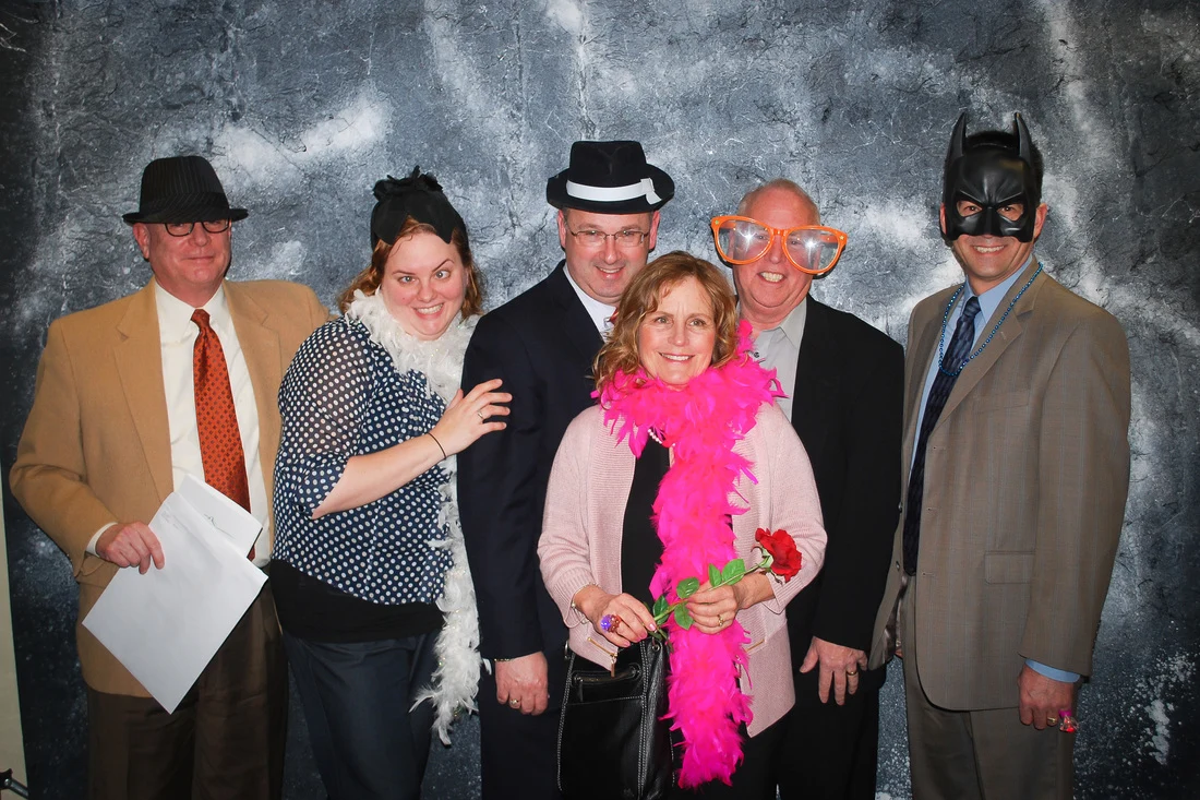 Six adults posing with party props, including hats, oversized glasses, a black mask, and a pink boa.