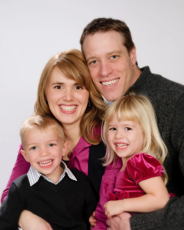 Two smiling adults and two children gathered closely together against a pale studio background.