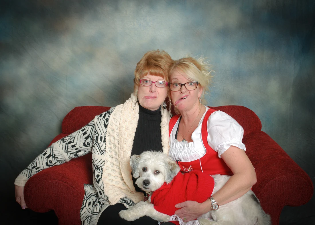 Two women with glasses sitting closely together on a red sofa and holding a small white dog.