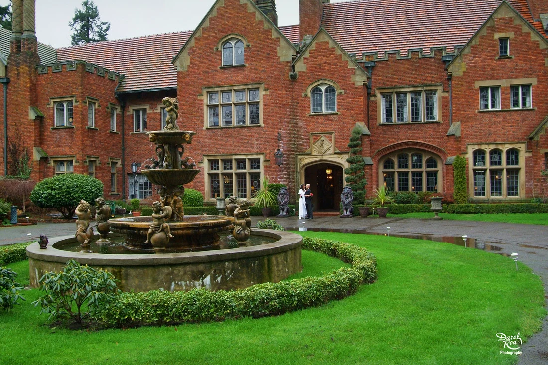 Bride and groom outside a red-brick manor, with a fountain and green lawn in the foreground.