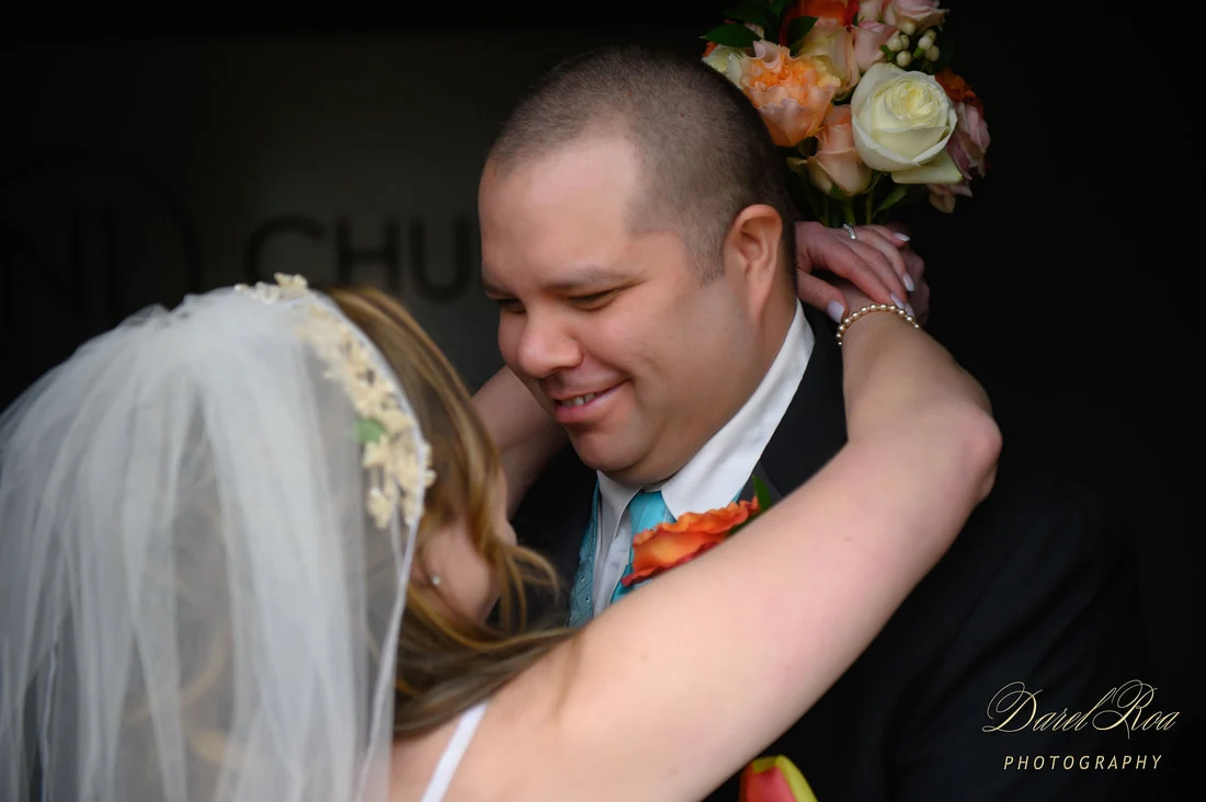 Bride embracing a smiling groom, with her veil and flower bouquet visible.